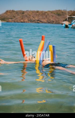 Eine Person genießt einen ruhigen Schwimmer mit bunten Poolnudeln im ruhigen blauen Wasser vor der Küste Costa Ricas, mit Booten und Hügeln in der Ferne Stockfoto