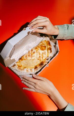 Ein frisch gebackener goldener Cheburek liegt in einer gemusterten Take-Out-Box, die von abgeschnittenen, nicht erkennbaren Frauenhänden gehalten wird, mit orange-rotem Hintergrund Stockfoto