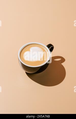 Von oben wunderschön gefertigter Cappuccino mit herzförmiger Latte Art, präsentiert in einem klassischen Keramikbecher mit weichem Schatten auf einem cremefarbenen Hintergrund Stockfoto