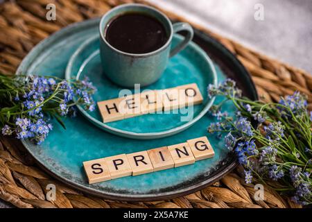 Ein gemütliches Kaffeeambiente mit einer Keramikbecher auf einem blaugrünen Teller, begleitet von lebhaften Vergissmeint-Not-Blumen und einladendem HELLO-FRÜHLING in Buchstaben Stockfoto