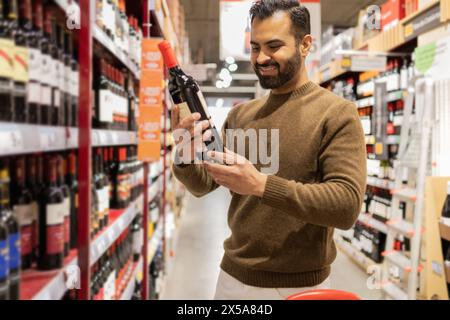 Lächelnder Mann, der eine Flasche Wein hält, während er in einem Supermarktgang shoppt Stockfoto