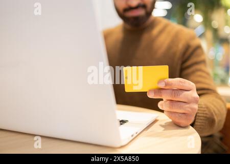 Nahaufnahme der Hand eines anonymen Mannes, der eine gelbe Kreditkarte vor einem Laptop hält und möglicherweise einen Online-Kauf tätigt Stockfoto