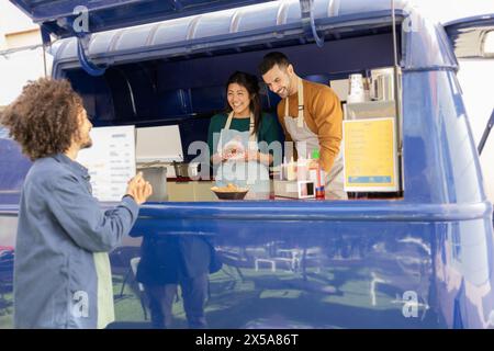 Fröhliche Freunde servieren einen zufriedenen Kunden aus ihrem leuchtenden, blauen Food Truck, der eine freundliche Street Food Business-Atmosphäre widerspiegelt Stockfoto