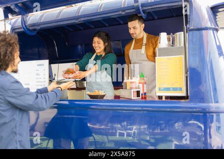 Fröhliche Freunde arbeiten in einem bunten Food Truck zusammen und überreichen einem zufriedenen Kunden eine köstliche Mahlzeit Stockfoto