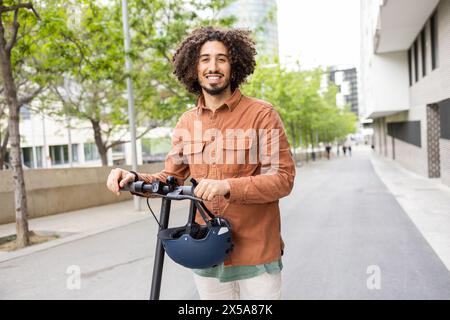 Selbstbewusster junger Mann mit lockigen Haaren, lächelnd, während er in einer modernen urbanen Umgebung mit einem elektrischen Roller fährt und einen Helm hält Stockfoto