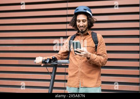 Ein fröhlicher junger Mann in einem Helm steht mit einem Elektroroller und greift sein Smartphone vor städtischer Kulisse an Stockfoto