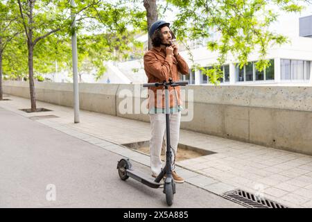 Ein junger Mann in lässiger Kleidung steht mit einem Elektroroller und hält sein Kinn nachdenklich in einem urbanen, von Bäumen gesäumten Bereich Stockfoto