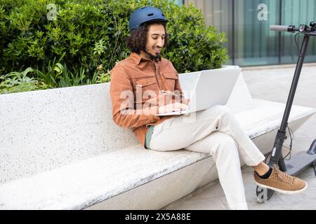 Ein junger Mann mit Helm sitzt entspannt auf einer Bank und arbeitet an einem Laptop neben seinem Elektroroller und verkörpert moderne urbane Mobilität und Technologie Stockfoto