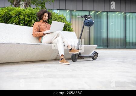 Ein junger Mann mit lockigen Haaren benutzt einen Laptop, während er auf einem modernen Elektroroller vor einem Gebäude mit Glasfassade sitzt, was eine Mischung aus Mobilität veranschaulicht Stockfoto