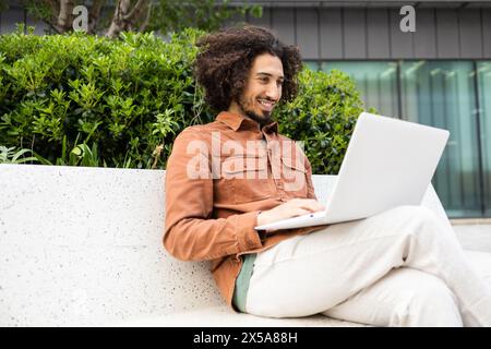 Ein Mann mit lockigen Haaren sitzt bequem auf einer Sitzbank im Freien, vertieft in seine Arbeit auf einem Laptop, umgeben von üppigem Grün, genießt die frische Luft während er die frische Luft genießt Stockfoto