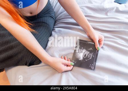 Eine schwangere Frau mit orangefarbenen Haaren sitzt auf einem Bett, hält zärtlich und sieht sich ein Ultraschallbild ihres ungeborenen Kindes an Stockfoto