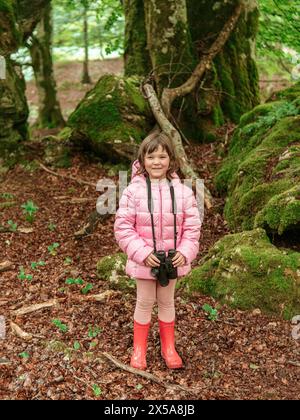 Ein junges Mädchen mit rosa Jacke und roten Stiefeln hält ein Fernglas in einem moosigen Wald und symbolisiert die Freizeit der Familie im Freien mit Großeltern. Stockfoto