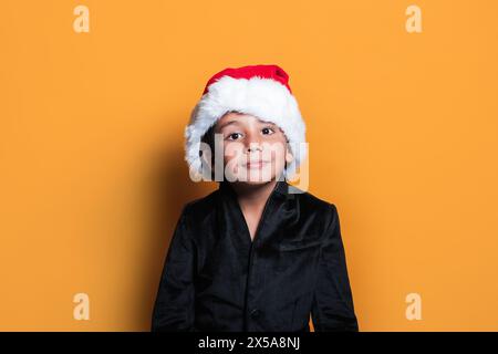 Ein kleines Kind mit festlichem Weihnachtsmann-Hut zaubert ein charmantes Lächeln in einem schwarzen Samtoutfit vor orangefarbenem Hintergrund Stockfoto