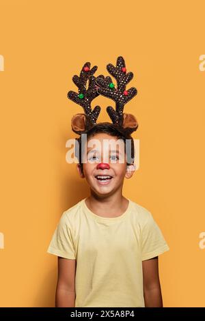 Ein fröhlicher kleiner Junge mit roter Nase und glitzerndem Rentiergeweih lacht vor einem warmen orangen Hintergrund und strahlt Feiertagsstimmung aus Stockfoto