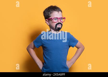 Ein kleines Kind steht selbstbewusst mit Händen auf den Hüften, trägt eine bunte Brille und einen aufgezogenen Bart vor einem leuchtend gelben Hintergrund in einem Studio-Set Stockfoto