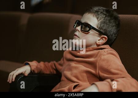 Ein kleiner Junge mit Brille schaut sich drinnen einen Film an, während er auf einem Sofa sitzt, was auf eine Freizeitaktivität im Sommer hindeutet. Stockfoto