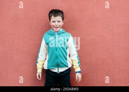 Ein glücklicher kleiner Junge steht selbstbewusst vor einer roten Wand in einer hellgrünen und gelben Jacke und genießt einen warmen Sommertag. Stockfoto