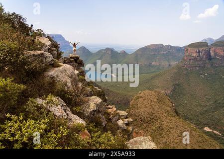 Rückansicht der unerkennbaren Frau steht auf einem Felsvorsprung mit erhobenen Armen und Blick auf den atemberaubenden Blyde River Canyon in Südafrika. Stockfoto