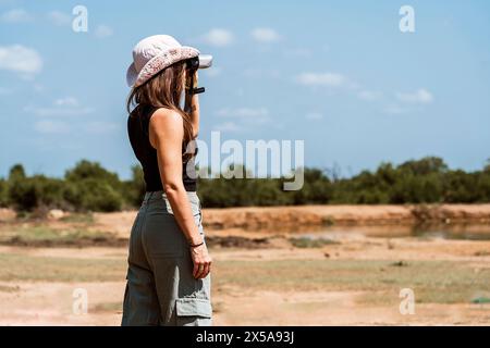 Seitenansicht einer nicht erkennbaren Naturliebhaberin mit Fernglas, gekleidet in Outdoor-Ausrüstung, Vogelbeobachtung in einer trockenen, grasbewachsenen afrikanischen Savanne. Stockfoto