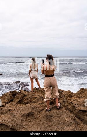 Rückansicht von zwei nicht erkennbaren Frauen stehen an einer zerklüfteten Küste, wobei eine mit einem Smartphone ein Foto vom Meer macht, während die andere aus dem Meer blickt Stockfoto