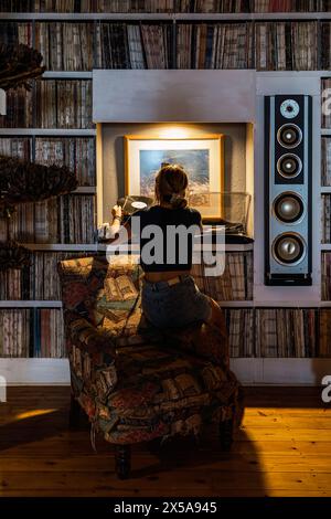 Rückansicht einer nicht erkennbaren Frau, die von hinten gesehen wird, während sie in einem Raum, der mit einer umfangreichen Vinylsammlung und einem Lautsprechersystem gefüllt ist, eine Schallplatte auflegt Stockfoto