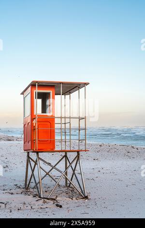 Ein leerer orangener Rettungsschwimmer-Turm steht an einem ruhigen Sandstrand vor einem klaren Himmel bei Sonnenaufgang, mit sanften Meereswellen im Hintergrund. Stockfoto