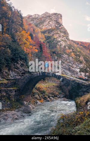 Rückansicht der nicht erkennbaren Person steht auf einer alten Steinbrücke über einem rauschenden Fluss, umgeben von den lebhaften Herbstfarben der Pyrenäen in Stockfoto