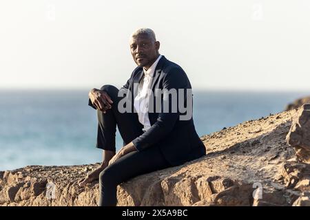 Afroamerikaner im Anzug sitzt nachdenklich in der Abenddämmerung auf den Felsen am Strand Stockfoto