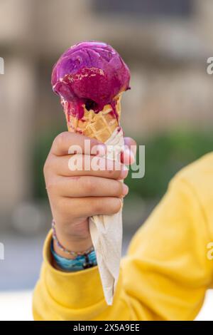 Bei einer Nahaufnahme wird eine leuchtende lila Eiskugel in einer Hand eines nicht erkennbaren Kindes gefangen gehalten, mit einer Papierserviette gewickelt und mit einer verschwommenen Hintergrundierung versehen Stockfoto