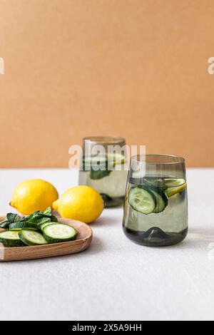 Zwei Gläser erfrischendes Gurken- und Zitronenwasser stehen neben frischen Zitronen und Gurkenscheiben auf einem Holztablett vor neutraler Kulisse. Stockfoto