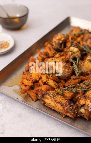 Gewürzte Hähnchenflügel neben Süßkartoffelwürfeln auf einem Tablett mit Dippsoße und Thymian bestreut. Stockfoto