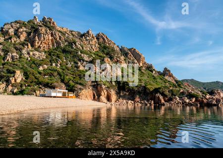 Ruhige Strandszene von Li Cossi in Paradiso Coast, Sardinien, mit zerklüfteten Hügeln und klarem blauen Himmel, der sich auf ruhigem Wasser spiegelt Stockfoto
