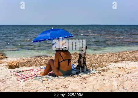 Eine Frau entspannt sich unter einem blauen Regenschirm mit ihrem treuen Hund an einem sonnigen Strand in Sardinien, Italien, und verkörpert den Geist der Sommerfreizeit Stockfoto