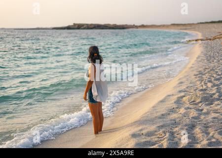 Eine Frau genießt einen friedlichen Spaziergang entlang eines Sandstrandes in Sardinien, Italien, während die Wellen sanft an der Küste während eines wunderschönen Sonnenuntergangs schlüpfen, ist Arutas Strand Stockfoto