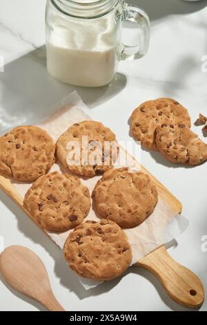 Köstliche Schokoladen-Chips-Cookies auf einem Backpapier neben einem Glas Milch und einem Holzlöffel, von oben unter natürlicher Beleuchtung aufgenommen Stockfoto