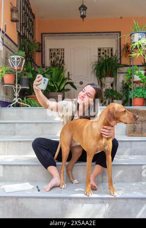 Eine lächelnde Frau macht ein Selfie mit ihrem goldenen Vizsla-Mischhund auf den Stufen eines Hauses mit Pflanzen und einer weißen Tür im Hintergrund Stockfoto