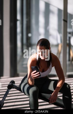 Eine fitte Frau in Sportbekleidung macht eine Pause vom Training zu Hause, um ihr Smartphone zu überprüfen und die Morgensonne zu genießen Stockfoto