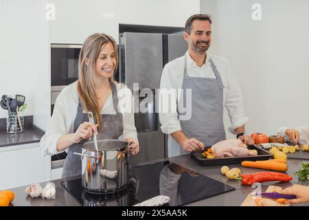 Eine fröhliche Familie verbindet sich, während sie ein köstliches gebratenes Huhn mit frischem Gemüse in einer modernen Küche kocht und dabei hausgemachte Gerichte vorführt Stockfoto