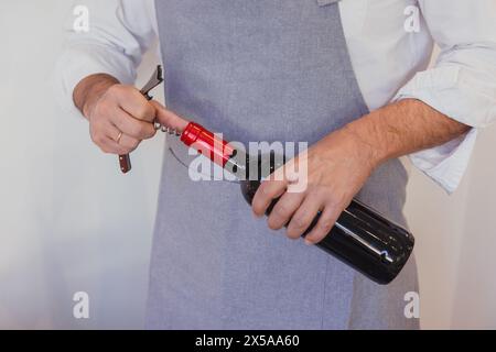 Anonyme Person in einer grauen Schürze entkorkt eine Rotweinflasche mit einem Korkenzieher Stockfoto