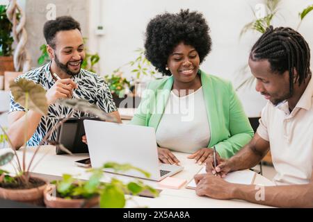 Ein dynamisches Team von drei Fachleuten nimmt an einer gemeinsamen Arbeitssitzung in einem hellen, mit Pflanzen gefüllten Coworking-Raum Teil Stockfoto