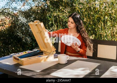 Eine Frau, die an einem Tisch sitzt, setzt sich mit ihren Malmaterialien in einer sonnigen Umgebung im Freien auseinander und ist bereit, ihre Kreativität auf Leinwand auszudrücken Stockfoto