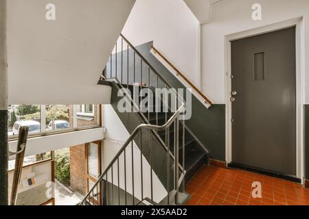 Eine gut beleuchtete Treppe in einem Wohngebäude mit Terrakotta-Fliesenboden und einem dunklen Metallgeländer führt zu einer grauen Tür. Stockfoto