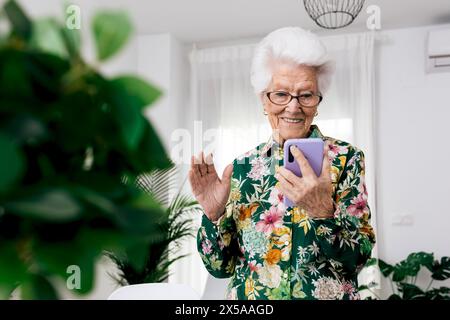 Lächelnde Seniorin in einem Blumenhemd, die in ihrem Wohnzimmer steht und glücklich winkt, während sie ein Smartphone hält Stockfoto