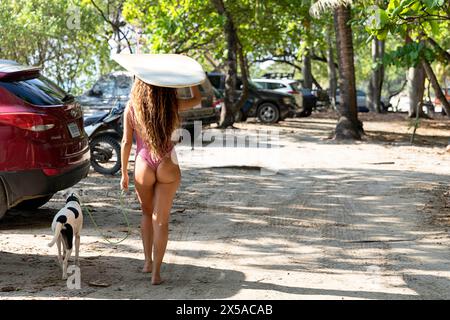 Eine Tourist, Frau im Badeanzug, die mit ihrem Hund und einem Surfbrett auf den Händen zum Sandstrand Santa Teresa auf der Nicoya-Halbinsel in Costa rica läuft Stockfoto