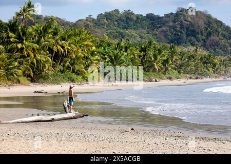 Kind, Tourist mit Angelrute auf einem Baumstamm auf einer Playa Hermosa, Strand auf Nicoya Halbinsel, mit Palmen und tropischem Wald auf der Rückseite, Costa Rica Stockfoto