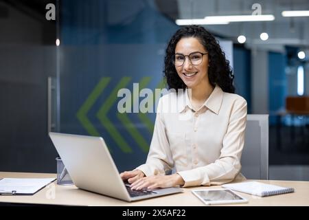 Junge, wunderschöne hispanische Frau, die in einem modernen Büro arbeitet, lächelt und mit einem Laptop in die Kamera schaut Stockfoto