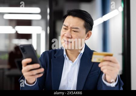 Ein asiatischer Geschäftsmann mittleren Alters sieht verwirrt und besorgt aus, während er seine Kreditkarte mit seinem Smartphone im Büro vergleicht. Konzept des Finanzbetrugs oder Bankfehlers. Stockfoto