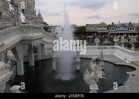 Ein Wasserbrunnen und Teich mit einer komplizierten und aufwendig geschnitzten Brücke und der Weiße Tempel in Thailand mit Touristen, die herumlaufen Stockfoto