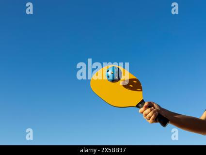 Arm, der einen Ball mit einem Paddel schlägt. Gelber Pickleball-Schläger, der einen blauen Ball trifft, mit Himmel im Hintergrund und Kopierraum Stockfoto