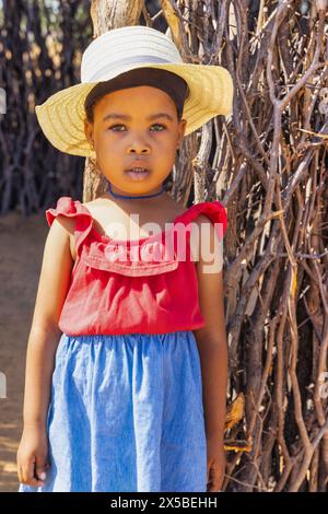 Afrikanisches Dorf, hübsches Mädchen, das vor der Außenküche im Hof steht, einen Strohhut und eine rote Bluse trägt Stockfoto